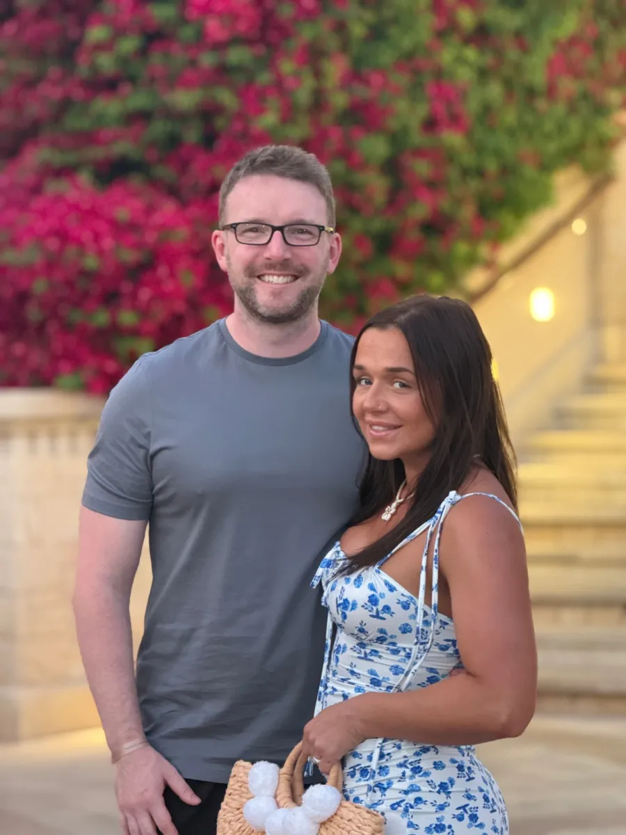 Shaun smiling beside his wife, in front of bright pink flowers
